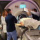 A patient surrounded by a medical team lays on the bed of an MRI machine with their head in a special focused ultrasound helmet
